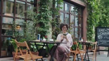 Kafe terasındaki masada oturan neşeli beyaz bir kadının gündüz vakti akıllı telefonuyla etrafa bakınması.