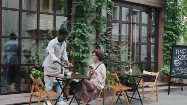 アフリカ系アメリカ人ウェイターのワイドショットが屋外テーブルに来て 白人女性にメニューを提供し 昼間に彼女の注文を書き留める — ストック動画