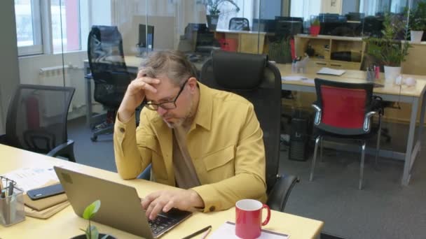 ノートパソコンの仕上げ作業でオフィスに座っている疲れた男性の白人マネージャーの中型ショット — ストック動画