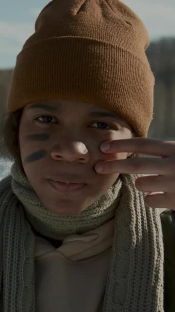 明るい冬の日に屋外に立っているウリーハットとスカーフで10代のアフリカ系アメリカ人の少女のクローズアップ垂直の肖像画ショット スノーボールが友人と戦う前に顔に戦争のストライプを描く — ストック動画
