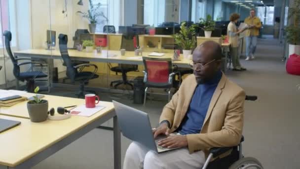 オープンスペースオフィスで膝の上にラップトップが付いている障害のある男性アフリカ系アメリカ人の従業員の北極の肖像画は笑顔で昼間カメラを見ています — ストック動画
