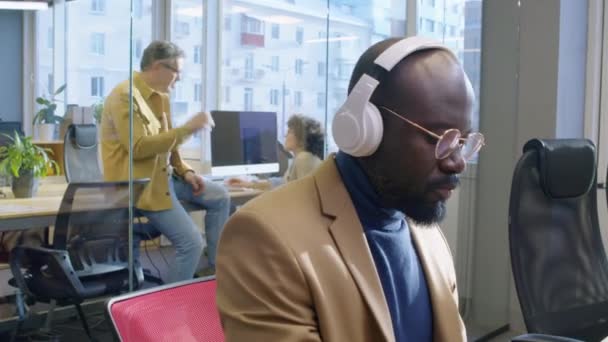 Muž Afroameričan Sedí Stolem Otevřené Kanceláři Průhlednými Stěnami Sluchátkách Pracuje — Stock video