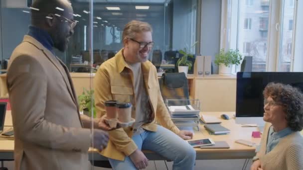 友好的なアフリカ系アメリカ人男性の右中型ショットを計画し 職場に座って話したり笑ったりする陽気な同僚2人にコーヒーをもたらす — ストック動画