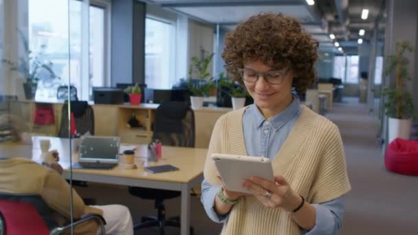 昼間タブレットで何かをしている間 オープンスペースオフィスに沿って歩いている白人女性の笑顔のスローモトラッキングショット — ストック動画