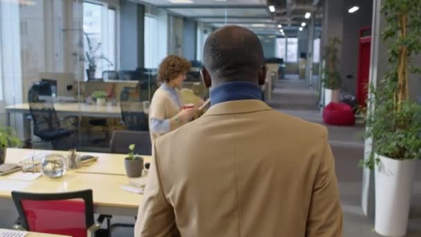 オフィスに沿って歩いているアフリカ系アメリカ人のマネージャーのスローモバックビュー追跡ショット 昼間働く多様な同僚 — ストック動画