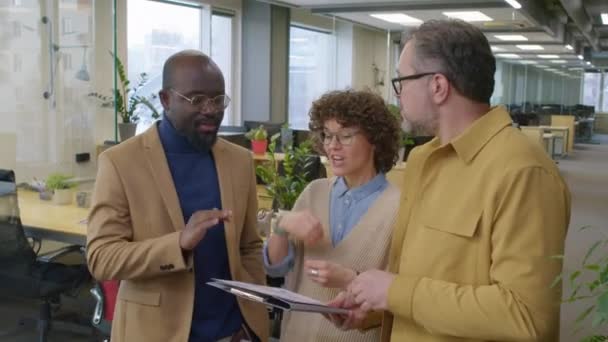 オフィスに立っている3人の多様な同僚の中型ショット 文書付きのフォルダを見て 昼間のプロジェクト戦略について議論 — ストック動画