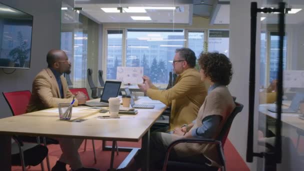 会議室のテーブルに座っている白人男性マネージャーの中央ショットは グラフとダイアグラムで示されたプロジェクト結果を昼間の多様な同僚に示しています — ストック動画