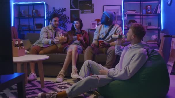 全景四照 四个衣着随意的年轻人在客厅的沙发上悠闲自在地躺着 点亮蓝色和粉色的霓虹灯 喝啤酒 分享薯片和聊天 — 图库视频影像