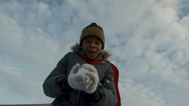 暖かいジャケットとウリーハットで9歳のアフリカ系アメリカ人の少年の中央低角ショット スノーボール カメラに向かって走り 友人のためのビデオメッセージを撮影し 遊ぶために呼び出します — ストック動画