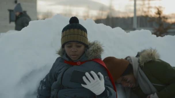 冬のジャケットで若いアフリカ系アメリカ人の男の子のミディアムショットと雪壁の背後でクロウチング 友人と一緒にスノーボールを投げ 楽しいソーシャルネットワークビデオを撮影 — ストック動画