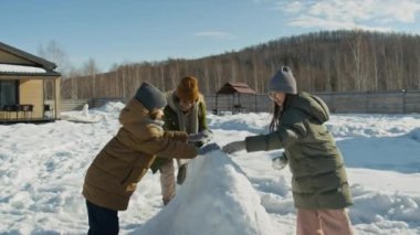 Güneşli bir günde arka bahçede oynayan ve birlikte kar heykeli yapan dört Afrikalı Amerikalı ve Kafkasyalı kız ve erkek çocuğunun orta boy fotoğrafı.