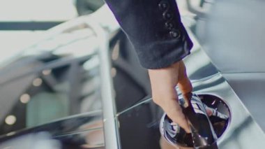 Orta boy dikey bir el resmi ve tanınmayan ceketli ve kazaklı genç adamın yansıması araba galerisinde veya garajda yeni siyah bir geçide doğru yürüyor, kapıyı açıyor ve içeri giriyor.