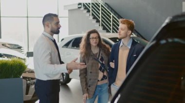 Çift ırklı bir satıcının galeride genç beyaz bir karı ve kocayla konuşurken orta kademe görüntüsü, araba özelliklerini ve özelliklerini açıklıyor ve araç modelleri öneriyor.