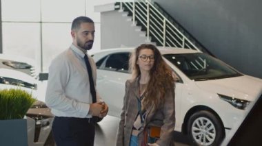 Orta boy, beyaz gömlekli ve kravatlı Orta Doğulu bir satıcının araba galerisinde günlük giysiler içinde genç beyaz bir kadınla konuşurken ve yeni çapraz model özelliklerini açıklarken.