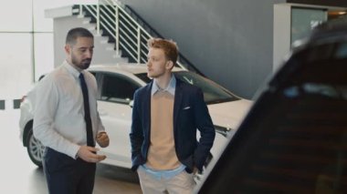 Orta boy, beyaz gömlekli ve kravatlı bir Orta Doğulu satıcının araba galerisinde günlük şık kıyafetler giyen beyaz iş adamıyla konuşması ve yeni çapraz modelin özelliklerini açıklaması.