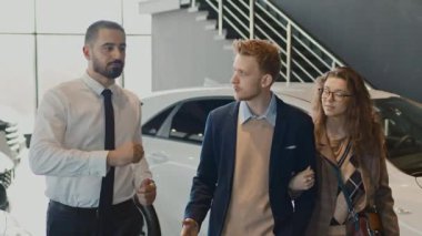 Genç karı ve kocanın otomobil galerisinde yürürken, farklı arabalara bakarken, beyaz gömlekli ve kravatlı bir satıcının yeni modeller için onlara danışmanlık yaparken orta boy çekimleri.