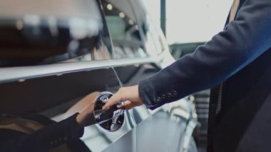 Ceketli ve süveterli, kimliği belirsiz genç bir erkeğin araba galerisindeki yeni siyah geçide doğru yürürken, kapıyı açarken ve test sürüşü için içeri girerken orta yavaş bir pozu.