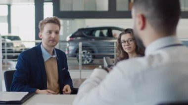 Otomobil galerisindeki satış müdürüne imzalı sözleşmeyi geri veren, elektronik anahtar alan, konuşan, dinleyen, gülümseyen ve gülümseyen genç bir adamın orta omuz üstü fotoğrafı.