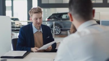 Orta boy, genç, beyaz bir karı kocanın araba galerisinde birlikte otururken, isimsiz bir temsilciyle konuşurken, oto satış sözleşmesinin kopyalarını okurken, şartları tartışırken.