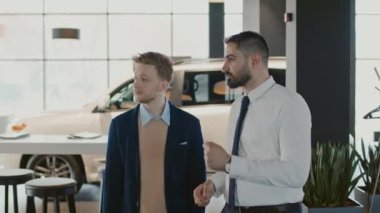 Orta boy Orta Doğulu genç satış müdürünün beyaz erkek müşteriyle otomobil galerisinde dolaşması, araba seçeneklerini ve özelliklerini tartışması ve vitrindeki yeni modellere işaret etmesi.