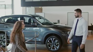 Orta ölçekli genç Orta Doğulu erkek satış temsilcisi araba galerisinde yürüyen, el sıkışan ve arabalara bakmaları için onları yönlendiren genç çifti selamlıyor.