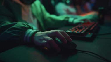 Yeşil bombardıman ceketli, bilgisayar faresini manipüle eden ve karanlık siber kafede atış oyunu oynarken klavye kullanan isimsiz bir gencin yakın plan fotoğrafları.