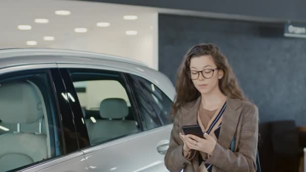 身着时髦休闲装 戴着眼镜的白人年轻女子在车行里走来走去 一边看智能手机 一边看正在展出的新的白色横线 — 图库视频影像