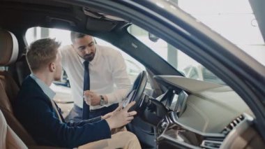 Sergi salonunda yeni arabanın ön koltuğunda oturan, sorular soran orta boy beyaz bir adamın orta boy fotoğrafı ve Orta Doğu satış temsilcisi, gösteriş paneli ve otomatik vites kutusu gösteriyor.