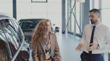 Orta halli Orta Doğulu bir satıcının genç beyaz kadın alıcıyla araba galerisinden geçerken, konuşurken, ön kapıyı açarken ve direksiyonun başına geçerken çekilmiş bir fotoğrafı.