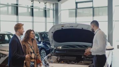 Beyaz gömlekli ve kravatlı genç bir Arap satıcının araba galerisinde kaputu açık bir şekilde duran ve genç karı-kocaya motor özellikleri hakkında konuşan yeni bir arabanın yanında durduğu orta boy fotoğraf.