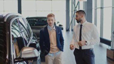Orta boy Orta Doğulu satış müdürünün beyaz erkek müşteriyle araba galerisinde yürürken görüntüsü. Konuşurken, ön kapıyı açarken ve direksiyonun başına geçerken.