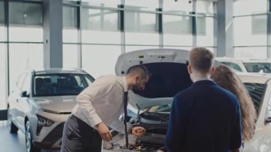 Erkek satış müdürünün açık kapüşonlu yeni beyaz arabanın yanında durup motor özelliklerini ve genç karı kocanın sorularını cevaplarken orta boy fotoğrafı.