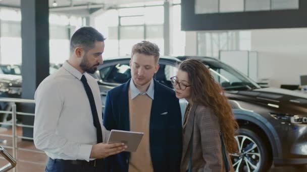 Średnie Ujęcie Sprzedawcy Samochodów Stojącego Salonie Rozmawiającego Parą Klientów Obliczającego — Wideo stockowe
