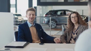 Beyaz tenli, şık ceketli, hırkalı, gözlüklü, sarı kıvırcık gözlü bir kadının araba galerisinde oturup satış müdürüyle sohbet ederken orta omuzlu bir fotoğrafı.