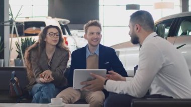 Genç beyaz bir çiftin araba galerisinde koltukta otururken orta boy fotoğrafı, Arap erkek satış müdürü tablet bilgisayarında mevcut araba renklerini gösteriyor, alıcılar tartışıyor, gülümsüyor ve kafa sallıyor.
