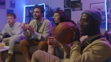 Orta boy beyaz bir kız ve erkek ve Orta Doğulu bir erkek arkadaşın kanepede oturup TV 'de spor maçı izlediği, ekranı gösterip tezahürat yaptığı ve siyahi bir adamın basketbolla oynadığı bir fotoğraf.