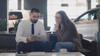 Genç beyaz kadın müşterinin otomobil galerisinde otururken orta boy fotoğrafı, elinde tabletle satış elemanının mevcut araba modellerini göstermesini, özellikleri ve seçenekleri tartışmasını,