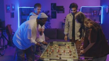 Orta boy bir grup öğrenci ev partisinde enerjik bir şekilde masa futbolu oynuyorlar, beyaz bir kız Afrikalı Amerikalı erkeği yeniyor, arkadaşlarıyla tezahürat yapıyor ve beşlik çakıyor.