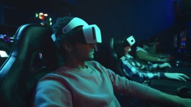 Siber kulüpte bilgisayar başında oturan, VR kulaklık takan, kafa sallayan, arttırılmış gerçeklikte birinci şahıs nişancı oyunu oynarken etrafa bakınan genç erkek oyuncunun orta boy yakın çekimi.