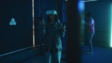 AR kulaklıklı, kot pantolonlu ve kot ceketli genç bir kızın karanlık odada neon ışıkla yürüdüğü ve el bilgisayarıyla sanal gerçeklik oyunu oynadığı orta boy fotoğraf.