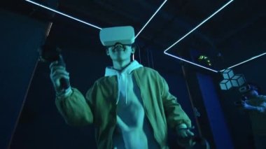 VR kulaklıklı genç adamın orta açılı görüntüsü. El kontrolörleri, boş karanlık odada, mavi neon ışıkta sanal gerçeklik savaşı oyunu oynuyorlar. Simülasyon nesneleri kesip geçiriyorlar.