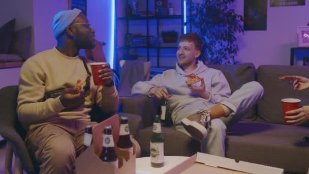 学生パーティーで友達と一緒に座って ピザを食べたり ビールを飲んだり 楽しい会話をしたり 笑ったりするアフリカ系アメリカ人と白人2人の若者のミディアムフルショット — ストック動画