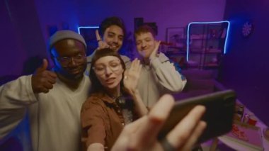Ev partisinde mavi neon ışıkta odanın ortasında duran, akıllı telefondan selfie çeken, komik yüzler ve boynuzlar yapan bir grup genç arkadaşın orta açılı görüntüsü.