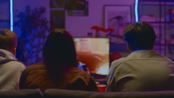 三个默默无闻的男孩和女孩在紫色霓虹灯下坐在沙发上 看着Fps游戏中的网络游戏在电视上流淌 并对失败嗤之以鼻 — 图库视频影像