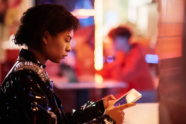 Young serious cyberpunk woman in shiny varnished jacket using tablet while sitting in front of camera in bar or cafe with neon lights