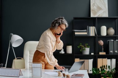 Akıllı telefonlu meşgul kadın patronun yan görünüşü. Masanın üzerine eğilip dizüstü bilgisayarda klavyeyle yazarken işi düzenliyor.