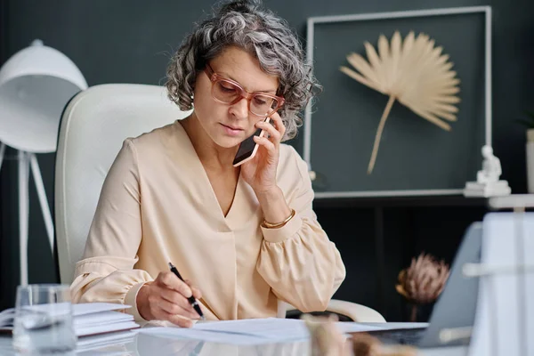 Confident mature woman in eyeglasses and quiet luxury attire phoning her subordinate and checking points of contract while sitting by desk