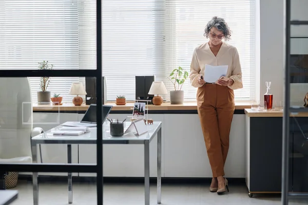 Mature female economist or broker in quiet luxury attire looking through financial paper standing by window covered with venetian blinds