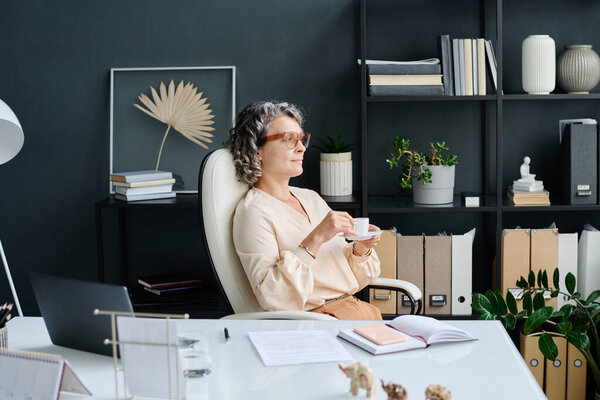 Restful mature female ceo with cup of coffee sitting by workplace in office, looking through window and thinking of something pleasant