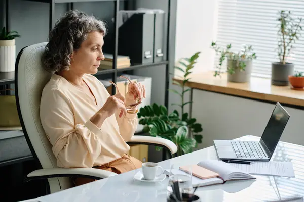 Serious female chief executive officer looking at laptop screen while sitting by desk in office and watching online video or reading information